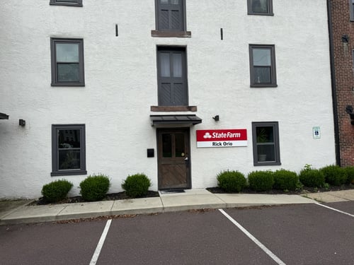 A white brick building with a parking lot in front and a red State Farm sign on the right side of the entrance door.