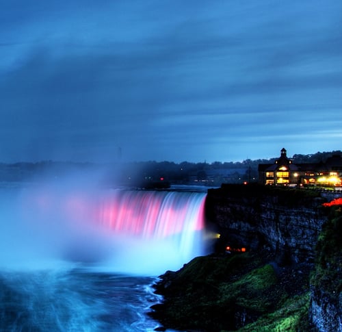 A vibrant waterfall cascades over a cliff at dusk, illuminated with pink lights. A building with glowing windows sits atop the cliff, creating a serene atmosphere.
