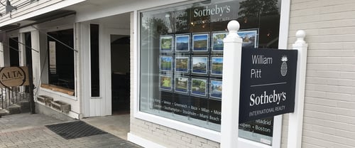 White building with covered entrance and large storefront window displaying property listings housing William Pitt Sotheby's International Realty in Lenox, Massachusetts, featuring decorative post with globe finial.