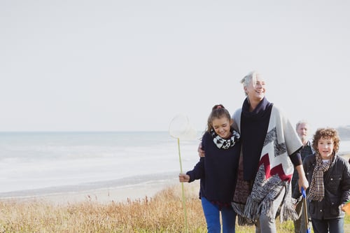 A joyful family strolls along a grassy beach path, with the ocean in the background. A young girl holds a fishing net, smiling alongside an older woman.