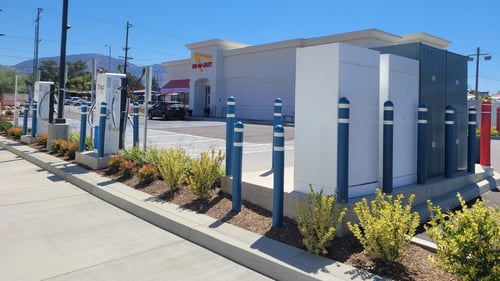 Photo taken from behind Several EVgo fast-charging units installed in marked parking spaces at a public charging station, with EVgo logos on the chargers, protective bollards around each unit