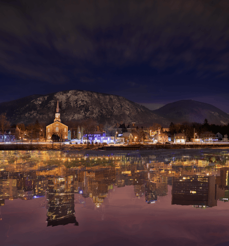 A serene night scene with a lit-up church and town buildings reflected in a calm river. Snow-capped mountains and a cloudy sky enhance the tranquil atmosphere.