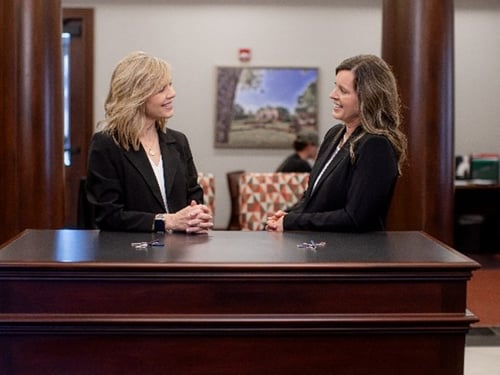 Two female Bryant Bank professionals in black blazers stand behind a polished wood counter, smiling and talking to one another. The background of the lobby features large wood columns and a framed landscape painting.