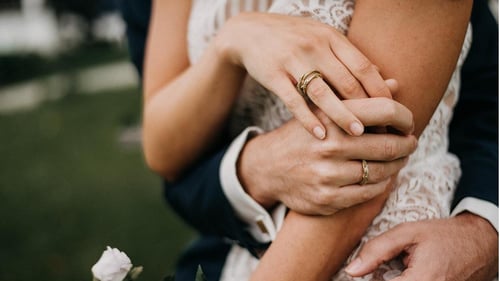 Ein Paar hält sich in den Armen, wobei beide ihre Hände zeigen, die mit handgefertigten goldenen Trauringen von Fingerglück, einem Schmuckgeschäft aus Stuttgart, geschmückt sind. Die Frau trägt ein Spitzenkleid, und im Vordergrund sind Blumen zu sehen. Die Szene vermittelt eine romantische Atmosphäre und zeigt die hochwertigen, handgefertigten Trauringe von Fingerglück.