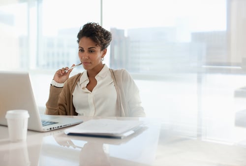 A woman in a white blouse and brown cardigan, holding a pen, thoughtfully looks at a laptop. A notebook and coffee cup are on her desk.