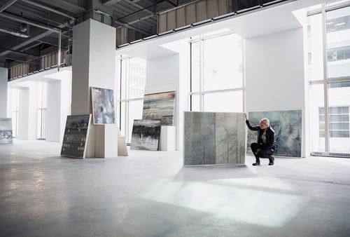 A person in a bright, spacious art gallery examines a large abstract painting. Sunlight streams through tall windows, illuminating several artworks.
