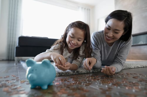 A woman and child lie on the floor, smiling as they count coins together. A blue piggy bank sits nearby, creating a warm, educational atmosphere.