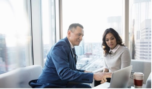 Business meeting in a modern office. A man in a blue suit and a woman in a sweater discuss work on a laptop. Bright cityscape view behind them.