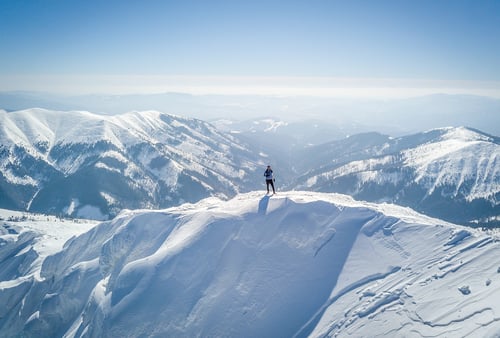 A lone skier stands on a snow-covered mountain peak, overlooking vast, snow-capped ranges under a clear blue sky, conveying a sense of solitude and adventure.