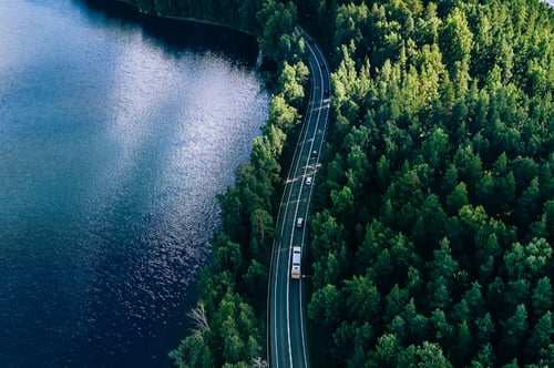 Aerial view of a winding road cutting through dense green forest beside a tranquil blue lake. Cars travel along the road, conveying a sense of peaceful journey.