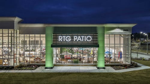 A night view of the RTG Patio storefront in Buford, GA, featuring bright green columns, floor-to-ceiling glass windows, and a clear view of the showroom’s furniture displays, creating an inviting atmosphere.