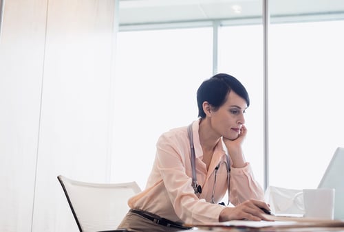 A doctor in a white coat sits at a desk, focused on a laptop, in a bright office. Stethoscope around neck; conveys concentration and professionalism.