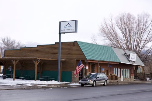 The exterior of an "Idaho First Bank" branch, which has a rustic, lodge-like appearance with a wooden facade and a green metal roof, under a cloudy sky. An American flag hangs by the entrance. A SUV is parked in front, and snow is on the ground.