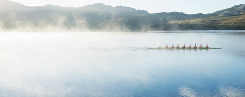 distant view of rowing team on still water approaching fog with mountain scape in the background.
