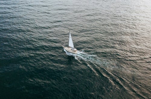 A lone sailboat glides through calm, rippling ocean waters, leaving a subtle wake behind. The scene conveys serenity and freedom on the open sea.