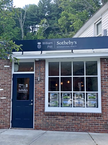 Brick building with blue door and large storefront windows housing William Pitt Sotheby's International Realty in Salisbury, Connecticut, featuring property listings display.
