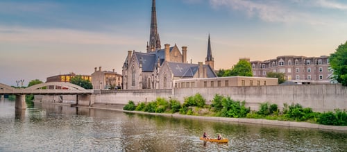 A historic church with a tall spire stands by a calm river. Two people in a yellow canoe paddle by, under a clear sky during sunset, exuding tranquility.
