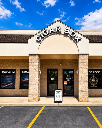 A well-maintained tobacconist storefront offering premium cigars, tobacco products, vape products, CBD products, and smoking accessories. The inviting entrance and convenient parking provide customers with a comfortable and easy shopping experience.