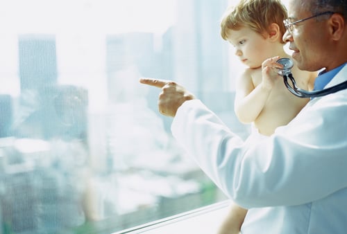 A doctor holds a stethoscope and points outside a window, holding a curious toddler. The image conveys care and curiosity in a bright setting.