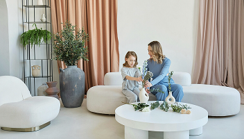 A woman and girl sit on a modern white sofa in a bright room with plants. They arrange flowers in vases on a round table, creating a calm, cozy atmosphere.