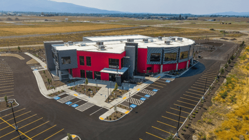 Peak Post Fall's new two-story gym has a striking red exterior and large windows. The building is surrounded by a well-maintained parking lot with ample spaces, including marked accessible spots. The gym is situated in an open area with expansive fields and mountains visible in the background.