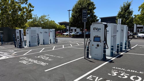Several EVgo fast charging units installed in marked parking spaces at a public charging station, with EVgo logos on the chargers, protective bollards around each unit.