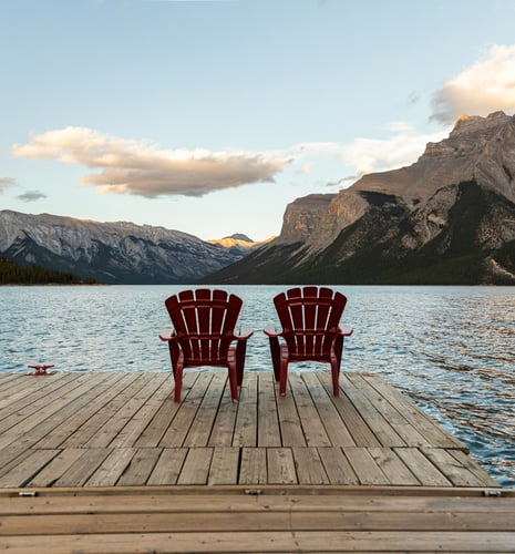Two red Muskoka chairs on a dock overlooking a lake with mountains in the background on a sunny day.