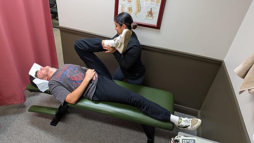 An assistant at Dr. Jonah Wassermann's office performing an assisted leg stretch and chiropractic adjustment on a male patient lying on a green treatment table, with a spinal column chart on the wall in the background.