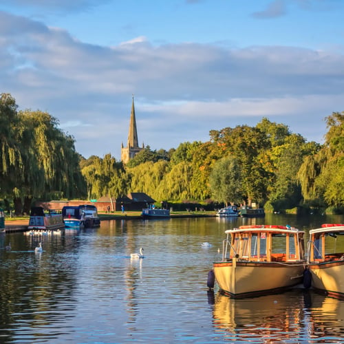 Knight Frank Stratford-upon-Avon Estate Agents - Scenic view of boats and swans on the River Avon with Holy Trinity Church spire in the background, Stratford-upon-Avon.