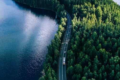 Aerial view of a winding road cutting through dense green forest beside a tranquil blue lake. Cars travel along the road, conveying a sense of peaceful journey.