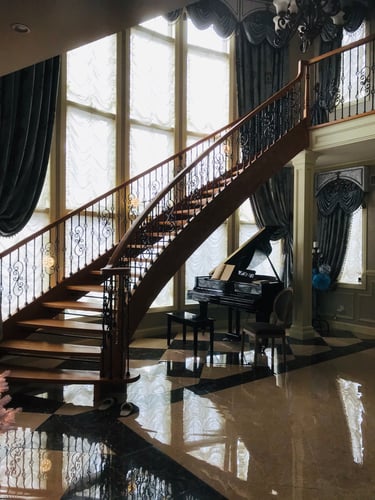 A spacious interior with a curved wooden staircase and ornate wrought-iron railings, tall windows draped with elegant curtains, a glossy geometric tiled floor, and a black grand piano beneath the staircase.