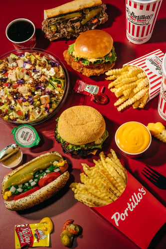A colorful overhead spread of Portillo’s menu items on a bright red background, featuring crinkle cut fries, a Chicago style hot dog, burgers, an Italian beef sandwich, a chopped salad, cheese sauce, drinks, and dipping sauces arranged around the table.