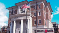 Exterior view of a Bryant Bank branch featuring a classic three-story brick building with prominent white Greek-style columns at the entrance. The American flag flies in front of the building under a clear blue sky.