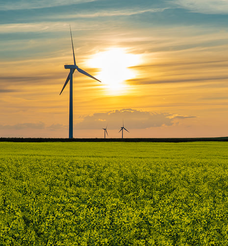 A serene landscape featuring two wind turbines silhouetted against a vibrant sunset. The foreground shows a lush, green field under a sky with golden hues.