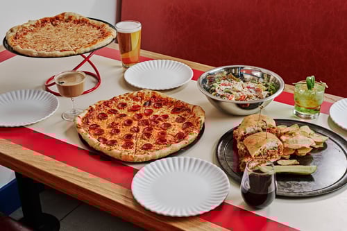 A table set with a large pepperoni pizza, a plain cheese pizza on a stand, a big salad, a hearty sandwich with chips and a pickle, white plates, and assorted drinks including beer, wine, and cocktails, suggesting a full dining experience.