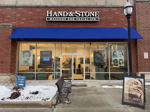 Exterior of Hand & Stone Lockport location with brick facade, blue awning, glass storefront, and promotional signage