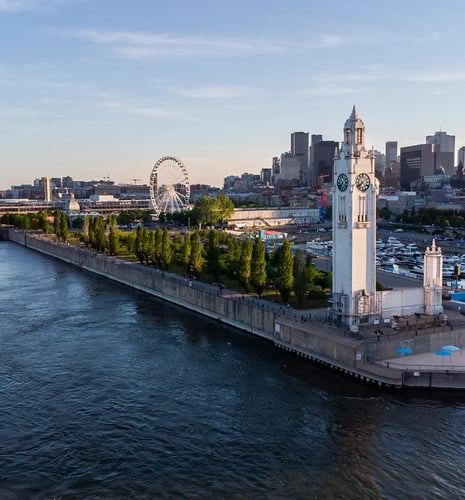 A scenic view of a city waterfront at sunset, featuring a prominent clock tower and lush green trees. A ferris wheel and city skyline are visible in the background.