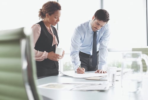 Business meeting scene with two professionals. A woman holds a mug, while a man in a tie writes on papers spread across the desk, conveying collaboration.