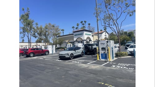 Several EVgo fast charging units installed in marked parking spaces at a public charging station, with EVgo logos on the chargers, protective bollards around each unit. NACS and CCS chargers featured in the image.