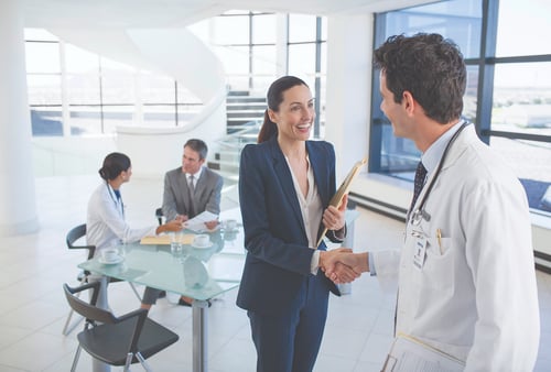 A doctor shakes hands with a smiling businesswoman holding a file in a bright office. In the background, two people converse at a glass table.