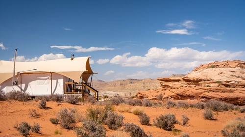 An Under Canvas Lake Powell glamping tent on one side with desert scenery, shrubs, and red-rock formations on the other.