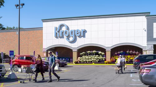 People with shopping carts walking through car lined parking lot in front of entrance of Kroger that has flowers in front