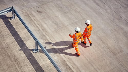 Aerial view of two construction workers in reflective orange clothing and hard hats walking on a concrete lot.