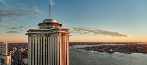 A tall building with a circular rooftop structure overlooks a winding river at sunset, with a distant cityscape and clouds illuminated by golden light.