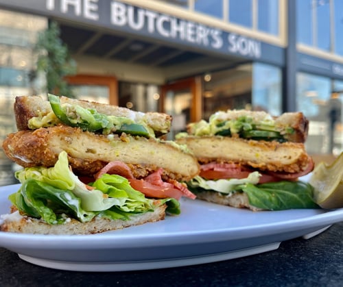 Plant-based killer BLAT (bacon, lettuce avocado, tomato) sandwich in front of The Butcher's Son vegan deli & bakery