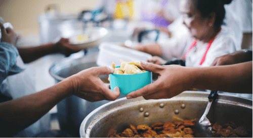 A close-up of hands passing a blue bowl filled with food at a busy soup kitchen. People in the background are serving meals. The scene conveys community and support.