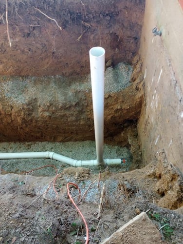 An open dirt trench with a newly installed white PVC drainage pipe, a vertical cleanout riser pipe, and a base layer of gravel bedding against a concrete foundation wall.