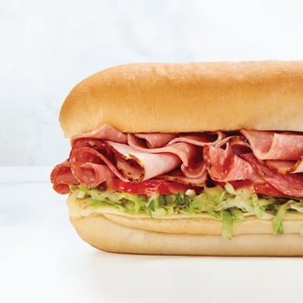 A close-up of a large submarine sandwich filled with layers of assorted deli meats, shredded lettuce, tomatoes, and sauce on a soft white sub roll.
