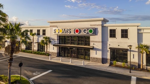 A front view of the Rooms To Go building featuring a clean white exterior, bold signage, palm trees, and large black-framed windows on a bright sunny day.