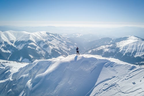 A lone skier stands on a snow-covered mountain peak, overlooking vast, snow-capped ranges under a clear blue sky, conveying a sense of solitude and adventure.